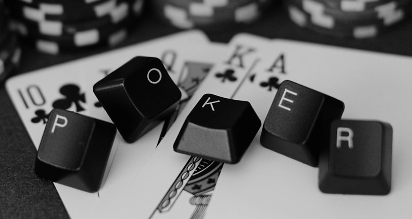 A series of playing cards and poker chips are displayed. The cards, including a ten, jack, queen, king, and ace of clubs, are spread out with black keyboard keys arranged to spell the word 'POKER'. The image is in black and white, giving it a classic, stylish look.