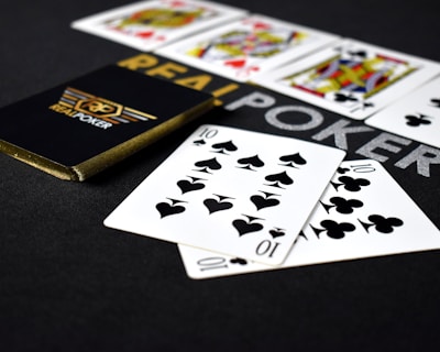 A deck of playing cards labeled 'Real Poker' rests on a dark surface. Two cards, the ten of spades and ten of clubs, are prominently displayed in the foreground. A row of face cards, including kings and queens from different suits, is lined up in the background.