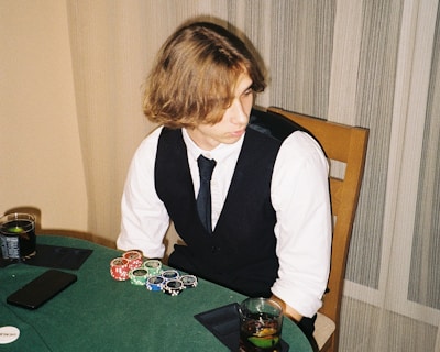 A person with shoulder-length hair is sitting at a poker table. They are wearing a white shirt, black vest, and tie, with an array of poker chips in front of them. There are two glasses with drinks on the table, and a smartphone is placed nearby. The background consists of vertical striped curtains.